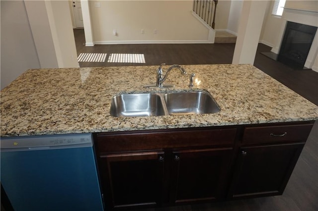 kitchen with a glass covered fireplace, dishwashing machine, light stone counters, dark brown cabinets, and a sink