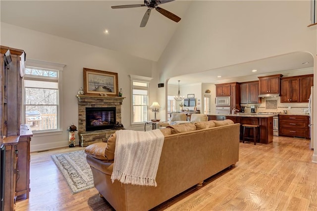 living area featuring ceiling fan with notable chandelier, a stone fireplace, light wood finished floors, and high vaulted ceiling