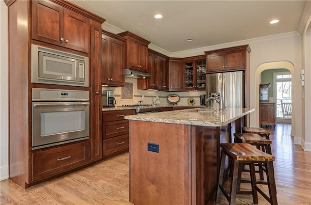 kitchen featuring a center island with sink, arched walkways, glass insert cabinets, stainless steel appliances, and a sink