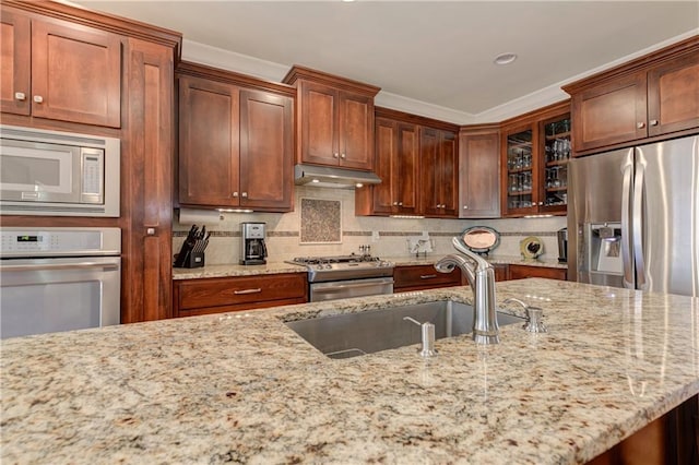 kitchen with glass insert cabinets, light stone countertops, stainless steel appliances, under cabinet range hood, and a sink