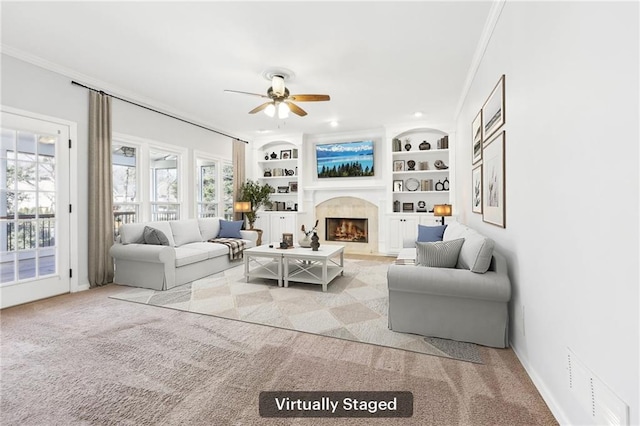 living area with ceiling fan, built in shelves, carpet flooring, a fireplace with flush hearth, and crown molding