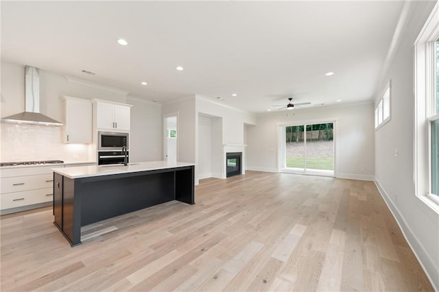 kitchen featuring stainless steel microwave, light wood-type flooring, wall chimney range hood, a kitchen island with sink, and wall oven