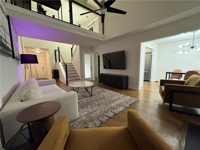 living room featuring stairway, a high ceiling, wood finished floors, baseboards, and ceiling fan with notable chandelier