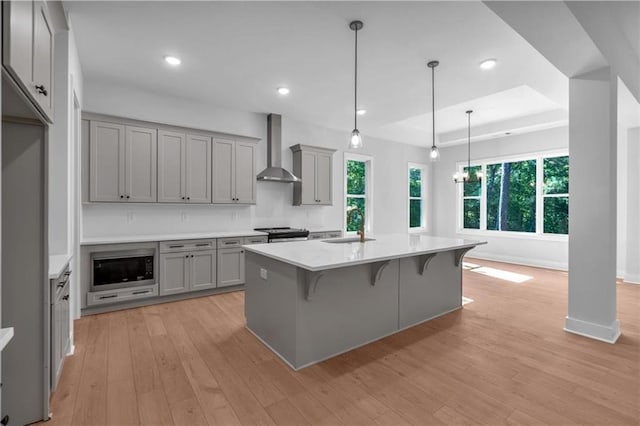 kitchen with gray cabinetry, wall chimney exhaust hood, a sink, light wood finished floors, and a raised ceiling