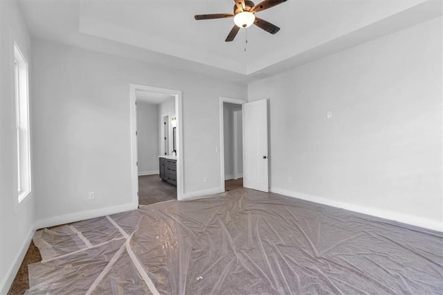 unfurnished bedroom with carpet, baseboards, a tray ceiling, connected bathroom, and ceiling fan