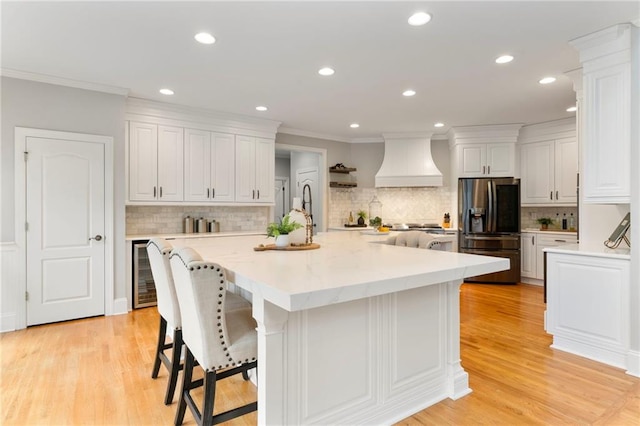 kitchen featuring white cabinets, black fridge with ice dispenser, wine cooler, ornamental molding, and premium range hood