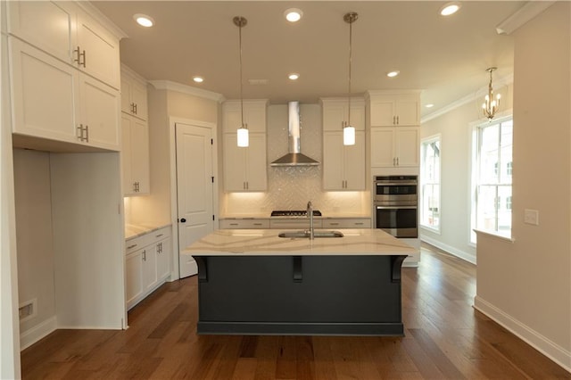 kitchen with light stone counters, hanging light fixtures, a center island with sink, stainless steel double oven, and wall chimney range hood