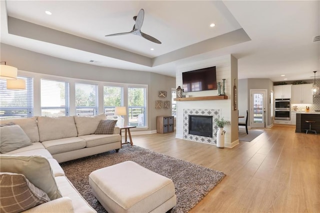 living room featuring a raised ceiling, light wood finished floors, baseboards, ceiling fan, and a tile fireplace