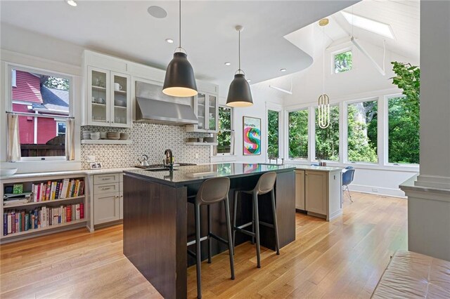 kitchen with wall chimney range hood, tasteful backsplash, a breakfast bar area, an island with sink, and light wood-type flooring