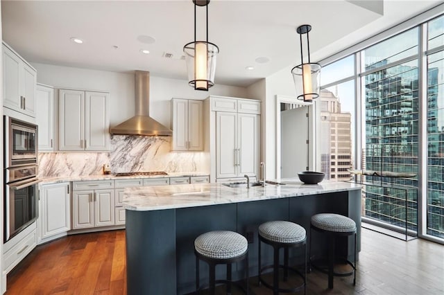 kitchen featuring appliances with stainless steel finishes, a kitchen island with sink, a wall of windows, wall chimney range hood, and white cabinetry