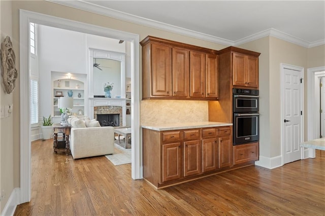 kitchen with wood finished floors, brown cabinetry, a stone fireplace, baseboards, and ornamental molding
