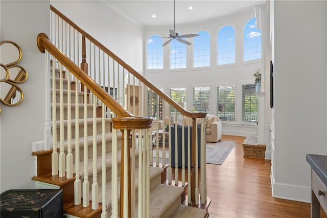 stairs with a ceiling fan, wood finished floors, a towering ceiling, baseboards, and crown molding