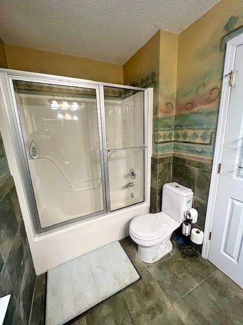 bathroom with a textured ceiling, toilet, tile walls, and combined bath / shower with glass door