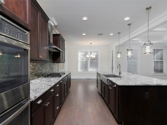 kitchen with wall chimney exhaust hood, an island with sink, decorative light fixtures, appliances with stainless steel finishes, and ornamental molding