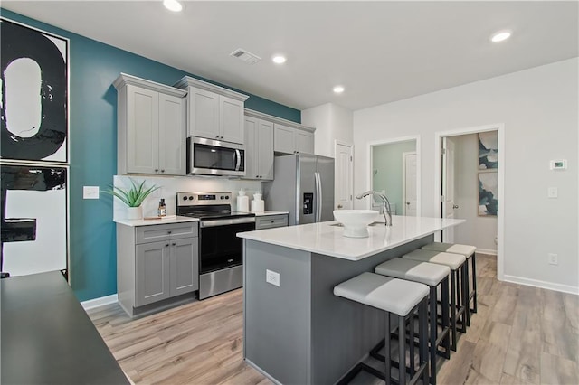 kitchen featuring light wood-type flooring, gray cabinetry, stainless steel appliances, a breakfast bar area, and an island with sink