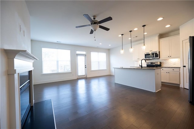 kitchen featuring a kitchen island with sink, white cabinetry, stainless steel appliances, decorative light fixtures, and dark stone counters