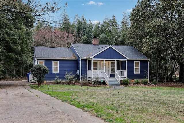 ranch-style house featuring a shingled roof, a chimney, covered porch, a front lawn, and board and batten siding