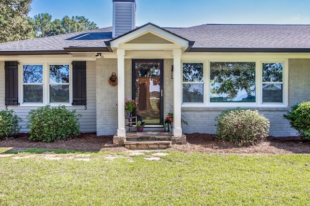 entrance to property featuring brick siding, a chimney, a lawn, and a shingled roof