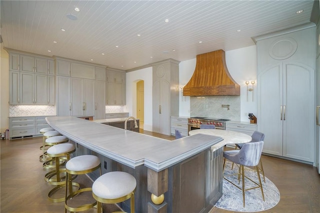 kitchen with high end stainless steel range oven, backsplash, a breakfast bar, an island with sink, and premium range hood