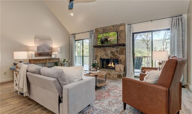 living room with ceiling fan, a stone fireplace, high vaulted ceiling, and light wood-type flooring