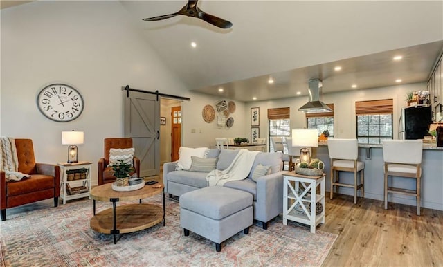 living room with ceiling fan, high vaulted ceiling, a barn door, and light hardwood / wood-style floors