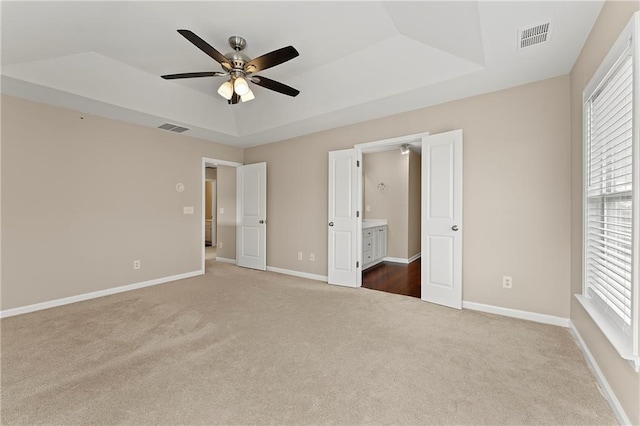 unfurnished bedroom featuring a tray ceiling, ensuite bath, ceiling fan, and carpet flooring