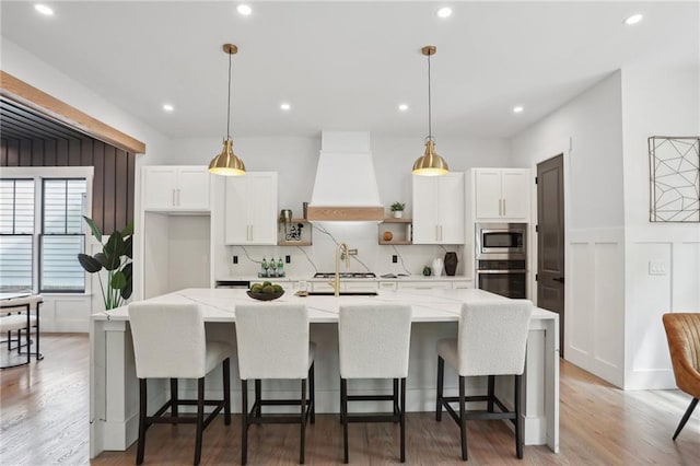 kitchen featuring hanging light fixtures, appliances with stainless steel finishes, a center island with sink, and premium range hood