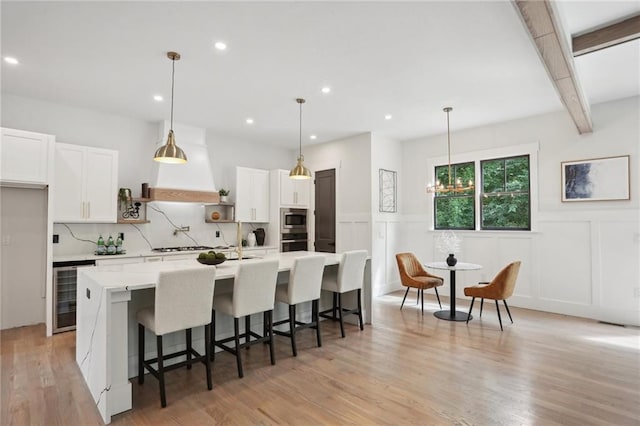 kitchen with a kitchen island with sink, beverage cooler, pendant lighting, beamed ceiling, and white cabinets