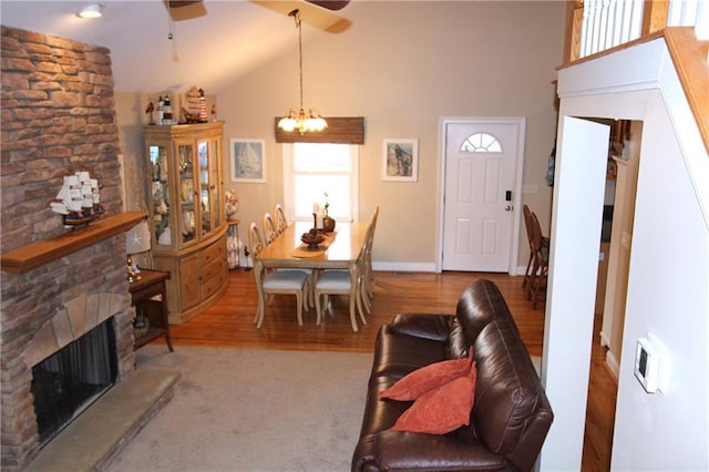 living area with ceiling fan with notable chandelier, high vaulted ceiling, a stone fireplace, and wood finished floors