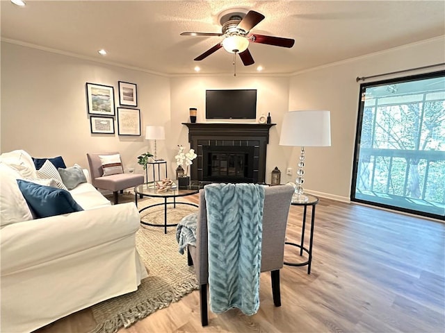 living area featuring baseboards, a ceiling fan, a glass covered fireplace, light wood-style flooring, and ornamental molding