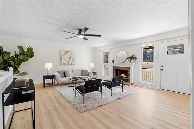 living room featuring a brick fireplace, ceiling fan, a textured ceiling, light wood-type flooring, and baseboards