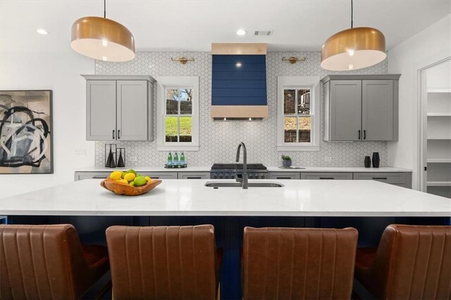 kitchen with gray cabinetry, backsplash, a center island with sink, and hanging light fixtures