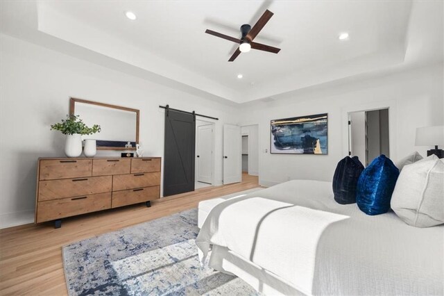 bedroom featuring ceiling fan, a tray ceiling, light hardwood / wood-style flooring, and a barn door