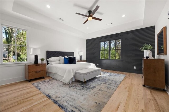 bedroom featuring ceiling fan, a tray ceiling, multiple windows, and light wood-type flooring