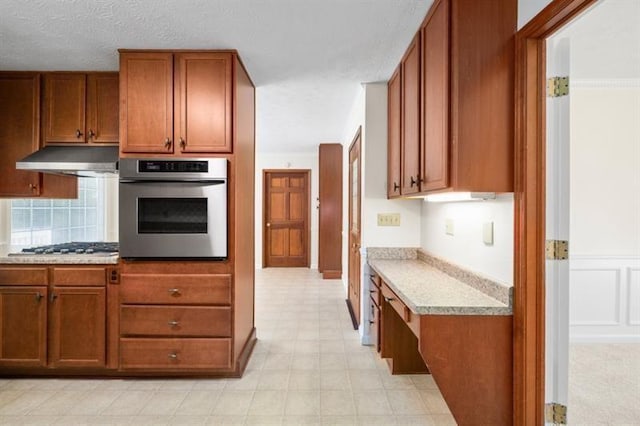 kitchen featuring under cabinet range hood, stainless steel appliances, light stone countertops, light floors, and brown cabinetry