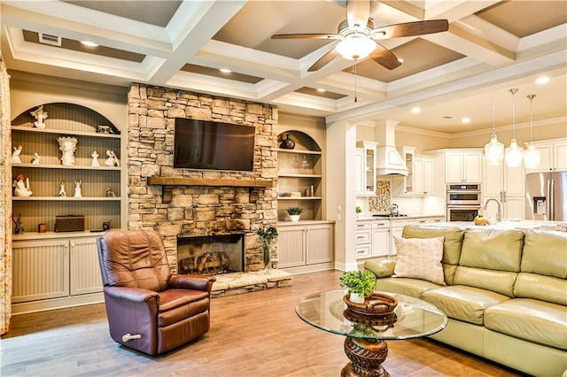 living room with coffered ceiling, a stone fireplace, crown molding, beamed ceiling, and light hardwood / wood-style floors