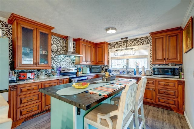 kitchen featuring stainless steel appliances, brown cabinets, wall chimney range hood, and wood finished floors