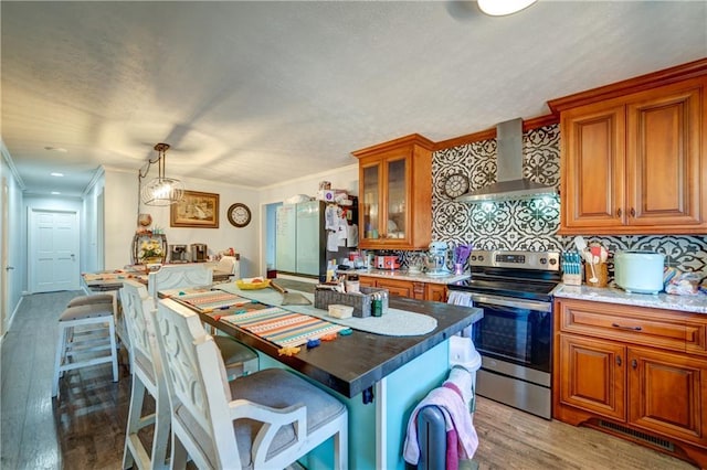 kitchen featuring crown molding, light wood finished floors, brown cabinetry, wall chimney range hood, and stainless steel electric range