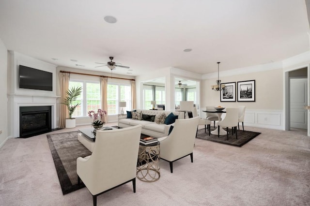 living area with light carpet, a decorative wall, ceiling fan with notable chandelier, a wainscoted wall, and a fireplace with flush hearth