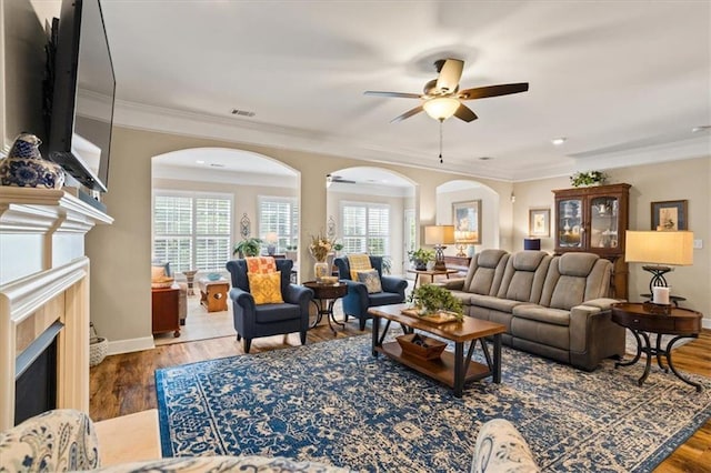 living area featuring a tile fireplace, ceiling fan, arched walkways, plenty of natural light, and crown molding