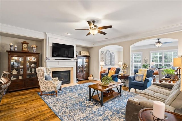 living room featuring a ceiling fan, a fireplace with flush hearth, ornamental molding, wood finished floors, and arched walkways