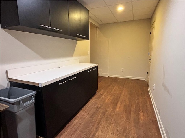 kitchen featuring dark cabinetry, dark wood finished floors, light countertops, a paneled ceiling, and baseboards