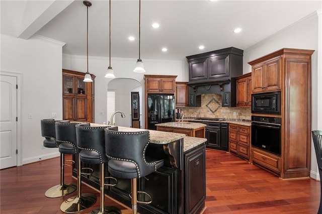 kitchen featuring dark wood-type flooring, an island with sink, light stone counters, pendant lighting, and black appliances