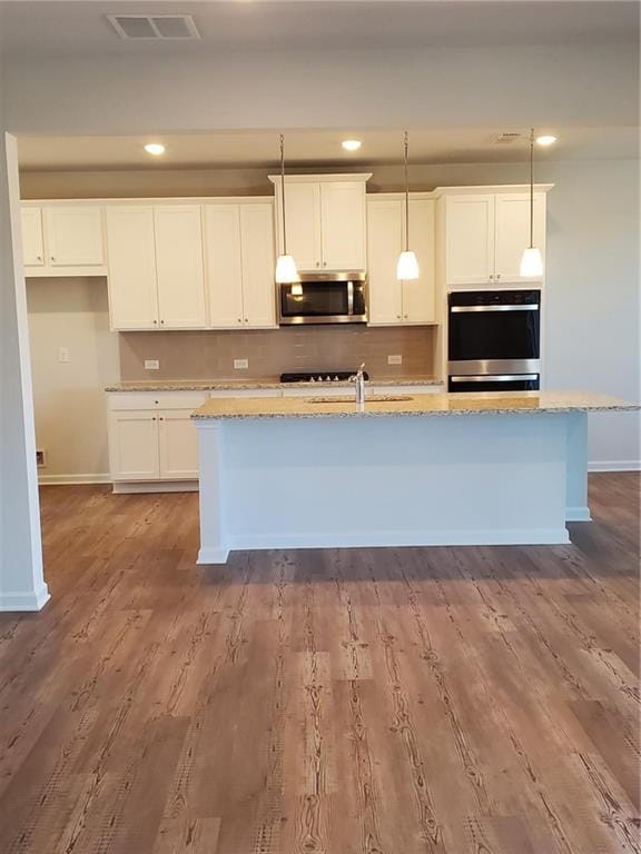 kitchen featuring an island with sink, stainless steel appliances, hanging light fixtures, and light stone countertops