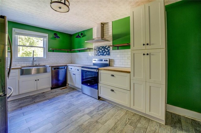 kitchen featuring wall chimney range hood, sink, light hardwood / wood-style floors, stainless steel appliances, and white cabinets