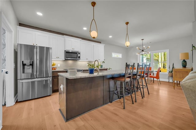 kitchen with hanging light fixtures, appliances with stainless steel finishes, a kitchen island with sink, white cabinets, and light wood-type flooring