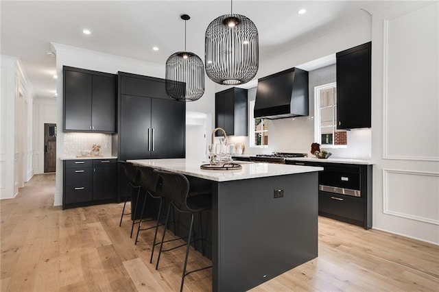 kitchen featuring custom exhaust hood, a kitchen island with sink, sink, light hardwood / wood-style flooring, and decorative light fixtures