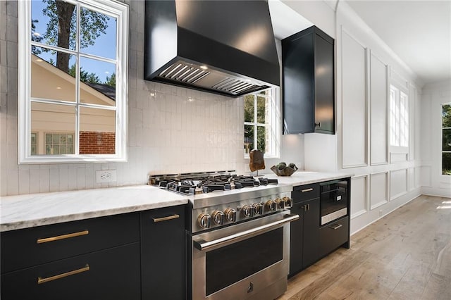 kitchen featuring wall chimney range hood, light stone countertops, high end stove, built in microwave, and light hardwood / wood-style floors