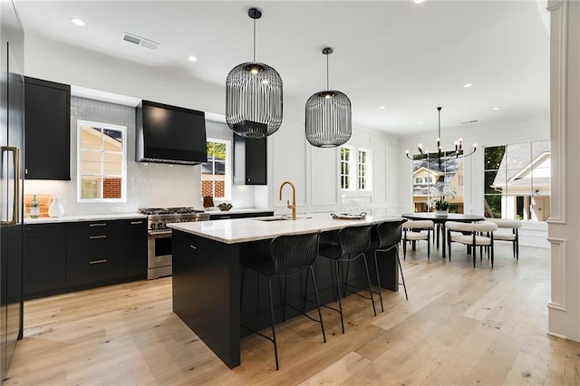 kitchen with exhaust hood, a center island with sink, stainless steel stove, light hardwood / wood-style floors, and hanging light fixtures