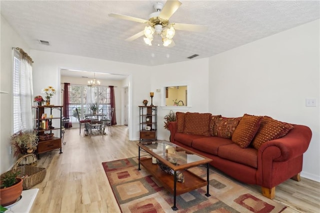 living area with a textured ceiling, ceiling fan with notable chandelier, visible vents, baseboards, and light wood finished floors